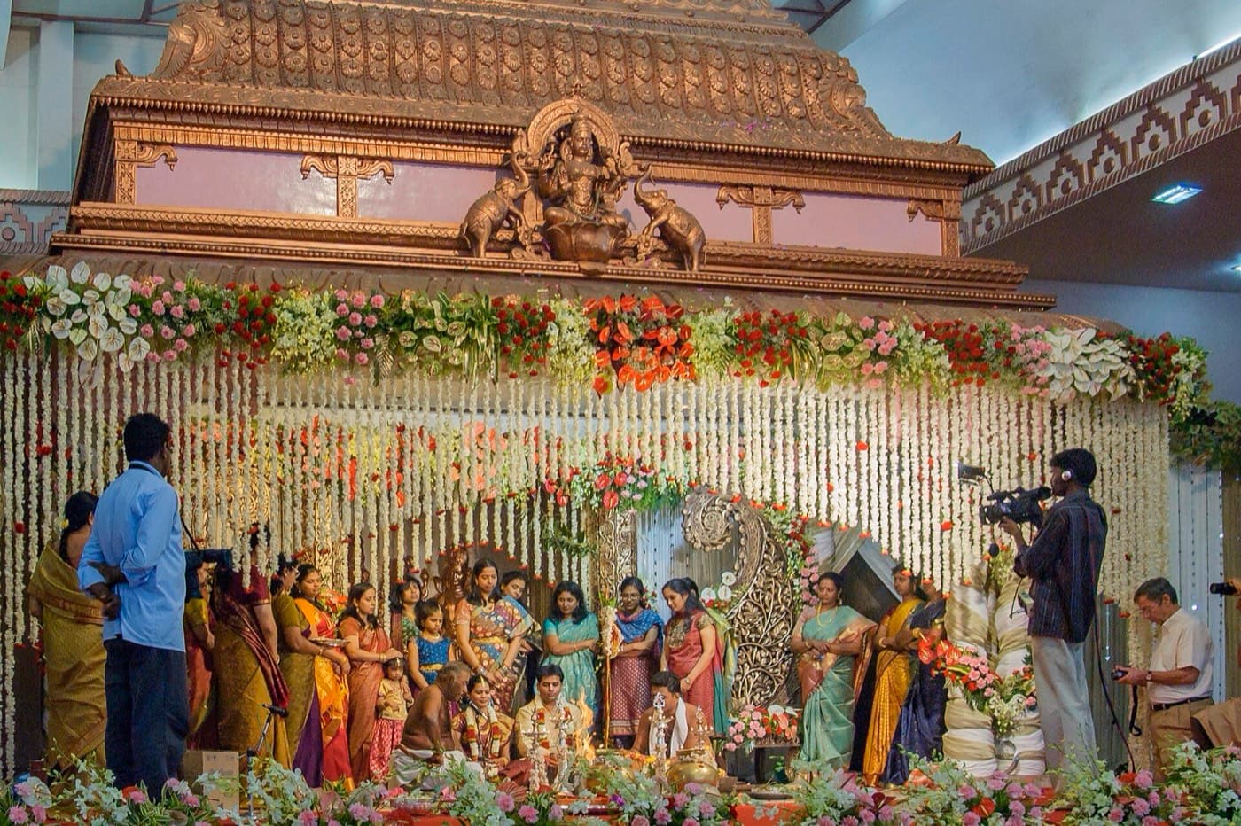A couple seated on a decorated South Indian wedding stage with jasmine garlands, surrounded by family