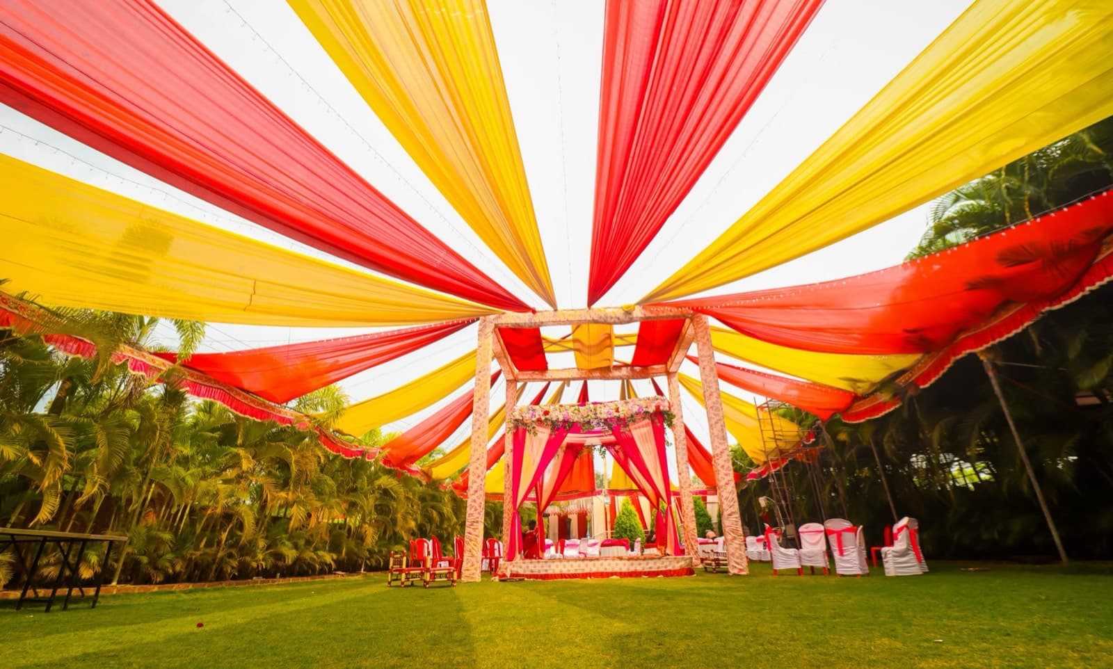 An outdoor wedding mandap under a red and yellow fabric canopy, with guest chairs on a lawn